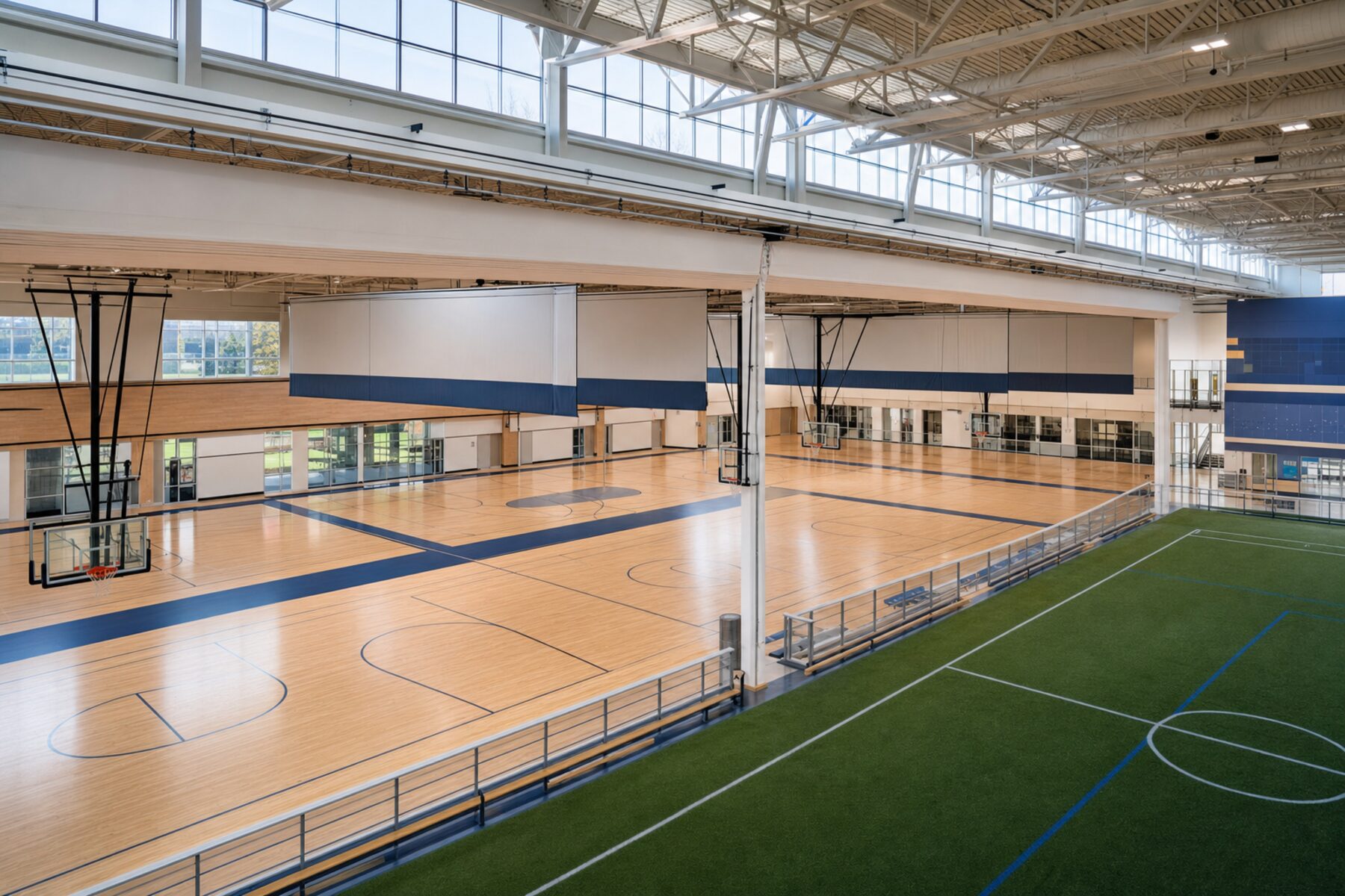 Bright indoor fieldhouse with divisible hardwood courts and adjacent turf under clerestory daylight